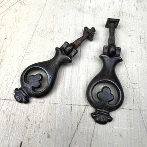 May include: Two antique black metal drawer pulls with a decorative floral design. Each pull features a teardrop shape with a central clover-like motif and a square-headed screw for mounting. The pulls are likely made of cast iron.