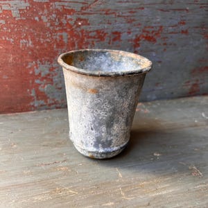 May include: A small, weathered metal cup with a slightly flared rim. The cup has a cylindrical shape and shows signs of rust and age, with a mottled gray and silver finish. The background is a distressed wooden surface with red and gray paint.