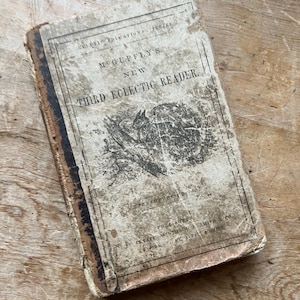 May include: An antique book titled "McGuffey's New Third Eclectic Reader" with a worn, distressed cover. The cover features text and a small illustration of an animal. The book is a vintage educational item.