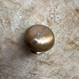 May include: A round, brass cabinet knob with a slightly oval shape. The knob has a smooth, polished finish and a subtle sheen.