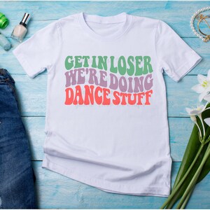 May include: White t-shirt with the text "Get in loser we're doing dance stuff" in a retro font with green, purple, and orange colors.