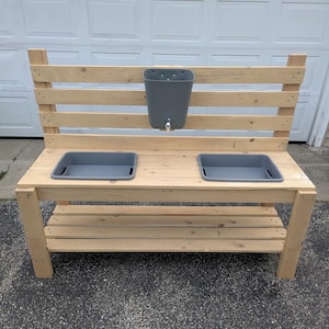 May include: A wooden outdoor play table with a backrest and a lower shelf. It features two gray plastic tubs, a gray plastic basin with a faucet, and is constructed from unfinished wood. The table is designed for children's outdoor activities.