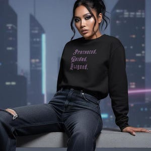 May include: Black sweatshirt with the words "Protected. Guided. Aligned." in a purple, gothic font. The model is wearing dark wash jeans with a distressed knee. The background features a blurred cityscape.