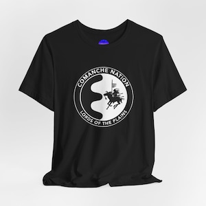 May include: Black t-shirt with a white graphic. The graphic is a circular design with the text "COMANCHE NATION" at the top and "LORDS OF THE PLAINS" at the bottom. Inside the circle is an image of a person on a horse.