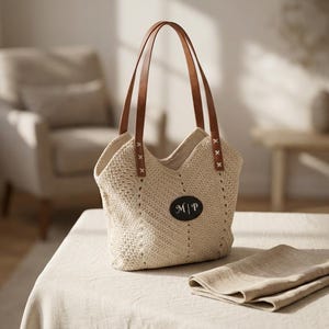 May include: A beige crochet tote bag featuring two brown leather handles attached with white cross-stitching. The bag has a central black oval patch with the white text M | P embroidered on it. The crochet pattern consists of diagonal lines.
