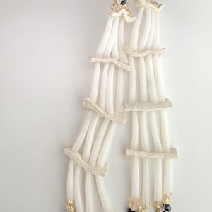 May include: White and cream-colored dangle earrings. The earrings feature multiple strands of white material, adorned with cream-colored lace accents and small black and gold beads. The earrings hang from silver-colored hooks.