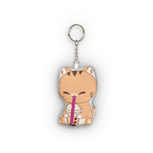 May include: A tan cat-shaped keychain with a silver keyring. The cat is smiling and drinking from a boba tea cup with a pink straw. The keychain is a soft, plush material.