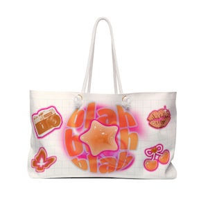 Puede incluir: Bolso tote blanco con un patrón de cuadrícula y un diseño con las palabras "blah blah blah" en naranja y rosa. El bolso tiene una estrella, labios, cerezas, una cámara y un gráfico de mariposa. Tiene asas de cuerda blanca.