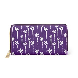 May include: A purple wallet with a white floral pattern and a gold zipper. The wallet is rectangular and has a zip-around closure. The floral design is a repeating pattern of white brushstrokes and flower shapes.