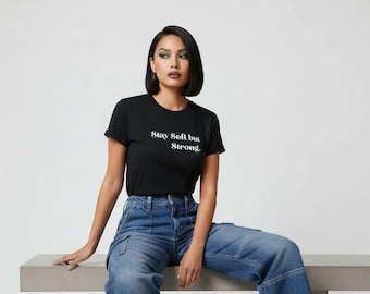 May include: Black crew neck t-shirt with the text "Stay Soft but Strong" in white. The model is wearing blue denim cargo jeans. The background is a plain white.