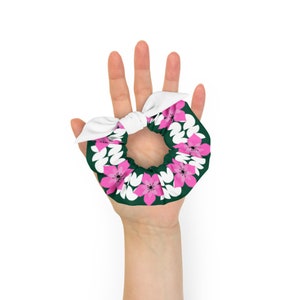 Puede incluir: Una scrunchie floral con un lazo blanco. La scrunchie presenta un fondo verde oscuro con un estampado de flores rosas y blancas. El lazo es blanco. Este accesorio para el cabello es perfecto para añadir un toque de color a cualquier peinado.