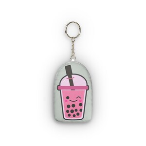 May include: A pink and black boba tea keychain with a smiling face. The keychain has a silver chain and ring. The boba tea cup has a pink lid and a black straw. The background is a light green color.