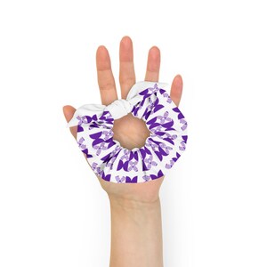 May include: A white scrunchie with a purple geometric pattern. The scrunchie is tied in a knot and is being held by a hand.