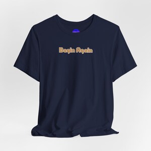 Könnte beinhalten: Marineblaues T-Shirt mit dem Text "Begin Again" in hellbrauner Schrift. Das Shirt hat kurze Ärmel und einen Rundhalsausschnitt. Der Stoff wirkt weich und leicht zerknittert.