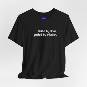 May include: Black t-shirt featuring the white text: "Ruled by tides, guided by intuition." The crew neck tee has short sleeves and a relaxed fit. The shirt is made of a soft material. The text is in a simple, sans-serif font.