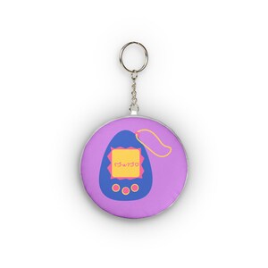 Puede incluir: Un llavero morado con un diseño de juego portátil retro. La pantalla del juego es azul con una pantalla amarilla y texto rosa. El llavero tiene un anillo y una cadena plateados.