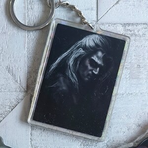 May include: A black and white image of a person's face with long hair is printed on a clear plastic keychain. The keychain has a silver metal ring and chain.