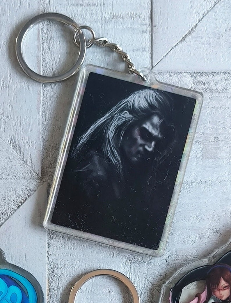May include: A black and white image of a person's face with long hair is printed on a clear plastic keychain. The keychain has a silver metal ring and chain.