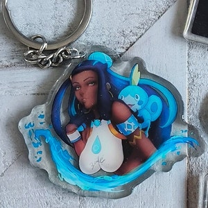 May include: A clear acrylic keychain featuring a blue-haired anime character with a blue water-type Pokemon. The character is wearing a white and gold outfit with a blue teardrop design. The keychain is surrounded by blue water-like swirls.
