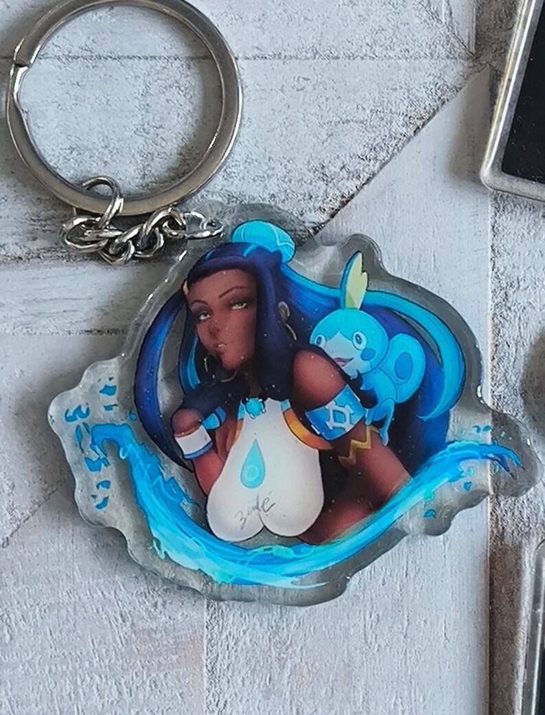 May include: A clear acrylic keychain featuring a blue-haired anime character with a blue water-type Pokemon. The character is wearing a white and gold outfit with a blue teardrop design. The keychain is surrounded by blue water-like swirls.