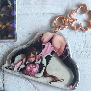 May include: A clear acrylic keychain featuring a cartoon anime character with long brown hair, wearing a pink and black outfit. The keychain has a gold metal clasp.