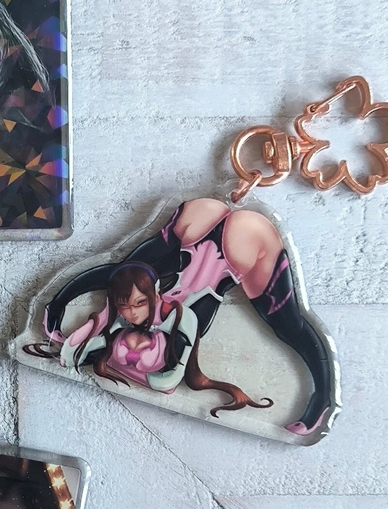 May include: A clear acrylic keychain featuring a cartoon anime character with long brown hair, wearing a pink and black outfit. The keychain has a gold metal clasp.