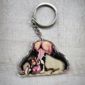 May include: A clear acrylic keychain featuring a colorful illustration of a woman in a pink and black outfit. The keychain is attached to a silver metal ring.