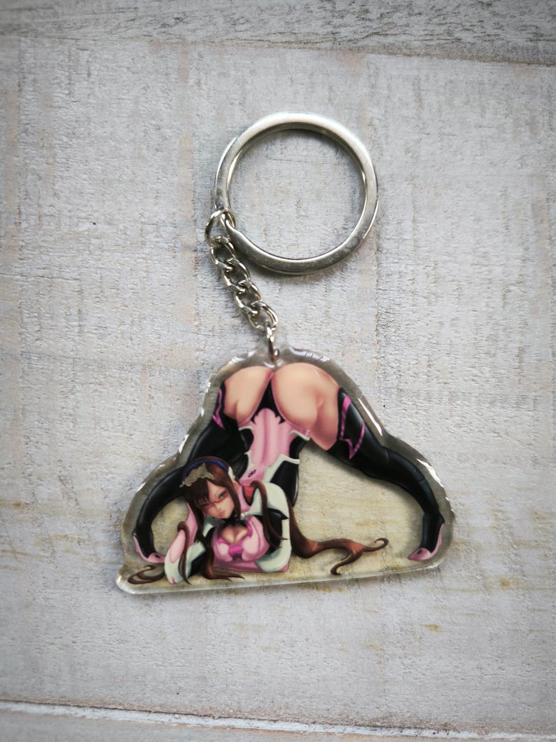 May include: A clear acrylic keychain featuring a colorful illustration of a woman in a pink and black outfit. The keychain is attached to a silver metal ring.