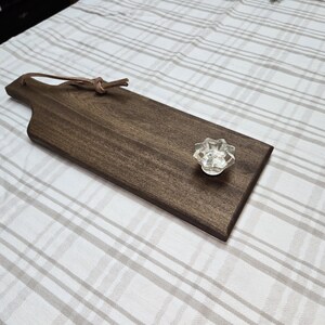 Puede incluir: Una tabla de servir de madera marrón oscuro con un asa de cuero y un pomo de vidrio transparente. La tabla está sobre un mantel a cuadros blanco y gris.