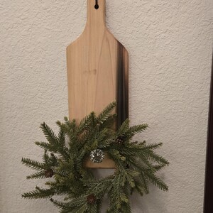 Puede incluir: Una tabla de cortar de madera con una mancha de madera oscura en la mitad inferior. Una pequeña corona de hoja perenne con piñas está colgada de la tabla.