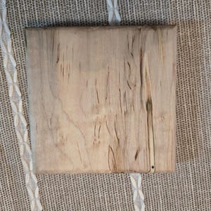 May include: A square, light-colored wooden board with visible grain and dark streaks. The board is set against a textured, neutral-toned fabric background with a white patterned border. The wood has a natural, rustic appearance.