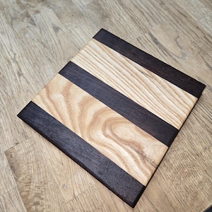 May include: A square wooden chopping board with alternating stripes of light and dark wood. The board is made of two different types of wood, one light and one dark.