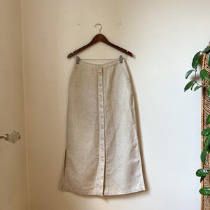May include: A beige linen button-down skirt hanging on a wooden hanger. The skirt has a high waist, a button closure down the front, and two front pockets. The skirt is knee-length with a front slit.