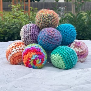 May include: A collection of nine colorful, crocheted balls in various shades of orange, blue, purple, green, and rainbow patterns. The balls are arranged on a white surface, with a blurred green background of foliage.