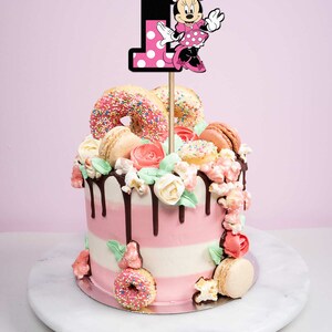 Pu&ograve; includere: Una torta a strati rosa e bianca decorata con ciambelle, macarons e fiori. In cima alla torta c'&egrave; un topper per torta di Minnie Mouse con il numero "1".