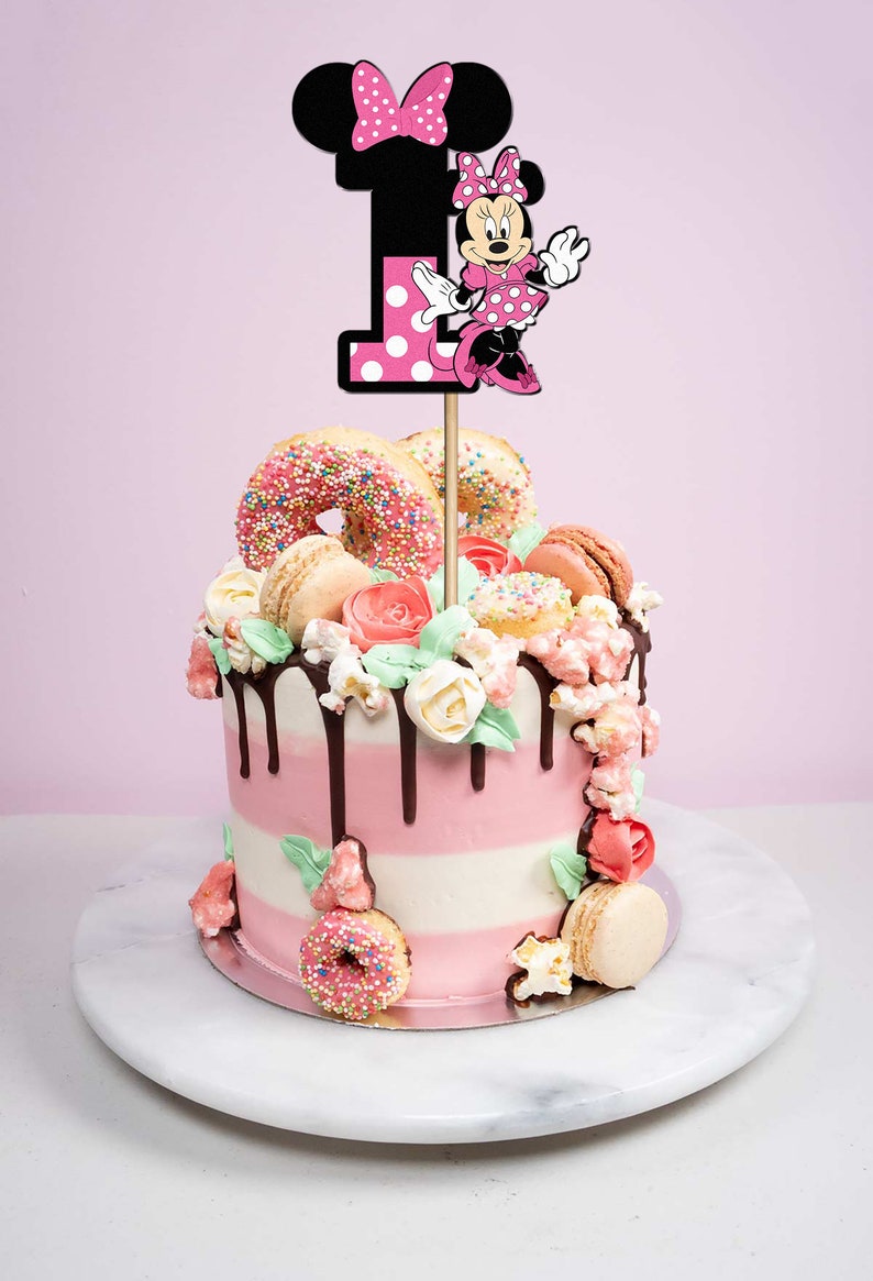 Pu&ograve; includere: Una torta a strati rosa e bianca decorata con ciambelle, macarons e fiori. In cima alla torta c'&egrave; un topper per torta di Minnie Mouse con il numero "1".