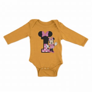Pu&ograve; includere: Un body per neonato a maniche lunghe color giallo senape con un numero uno a pois rosa e bianchi e un motivo di Minnie Mouse.