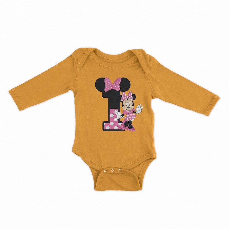 Pu&ograve; includere: Un body per neonato a maniche lunghe color giallo senape con un numero uno a pois rosa e bianchi e un motivo di Minnie Mouse.