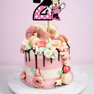 May include: A pink and white layered cake decorated with donuts, macarons, popcorn, and flowers. A black and pink Minnie Mouse topper with the number "2" is on top of the cake.