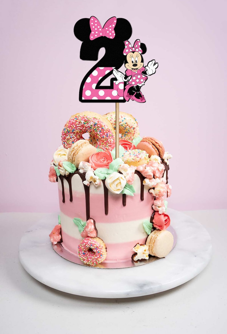May include: A pink and white layered cake decorated with donuts, macarons, popcorn, and flowers. A black and pink Minnie Mouse topper with the number "2" is on top of the cake.