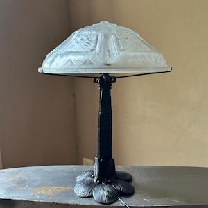 Può includere: Una lampada da tavolo con paralume a cupola in vetro smerigliato, decorato con motivi geometrici e floreali in rilievo. La lampada presenta uno stelo in metallo scuro e una base composta da petali testurizzati sovrapposti tra loro.