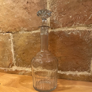 Puede incluir: Una decantador de vidrio transparente con un tapón facetado. La decantador tiene un cuerpo redondo con una superficie texturizada.