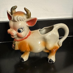 Puede incluir: Una lechera de cerámica con forma de vaca. La vaca es de color crema con detalles en naranja y marrón, un collar rojo y una campana. Tiene grandes ojos azules, orejas rosas y pequeños cuernos. La lechera tiene un asa y un pico vertedor.