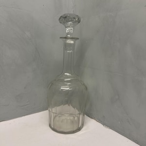 Puede incluir: Una decantador de vidrio transparente con un tapón de vidrio facetado. La decantador tiene un cuerpo redondeado y un cuello alto y delgado.