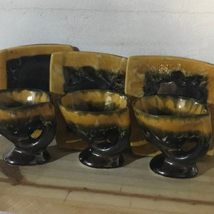 Peut inclure: Ensemble de trois bols en céramique avec des poignées en forme de mains. Les bols sont émaillés dans des tons de brun, jaune et noir. Chaque bol est associé à une assiette carrée assortie.