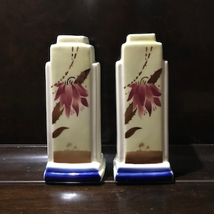 Puede incluir: Un par de jarrones de cerámica blanca con base azul y diseño floral. Los jarrones presentan una flor rosa con hojas marrones y un fondo marrón.