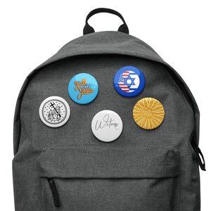 May include: A gray backpack with five different button pins attached. The pins feature a variety of designs, including a cross with a lamb, a blue pin with the text "Y'all need Jesus", a white pin with the text "Witness", a pin with the American and Israeli flags, and a gold pin with a sunburst design.