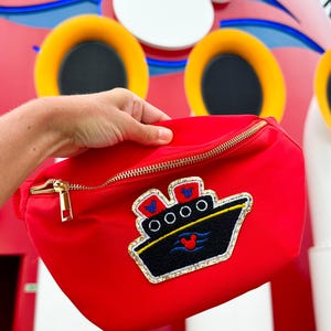 May include: A bright red fanny pack featuring a gold-tone zipper and an embroidered patch of a black cruise ship. The ship patch includes two red smokestacks with blue Mickey Mouse icons, white portholes, and a red Mickey Mouse icon on the hull.