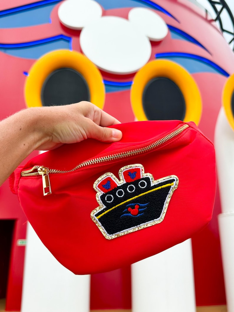 May include: A bright red fanny pack featuring a gold-tone zipper and an embroidered patch of a black cruise ship. The ship patch includes two red smokestacks with blue Mickey Mouse icons, white portholes, and a red Mickey Mouse icon on the hull.