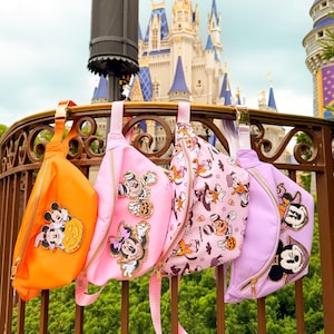 May include: Four fanny packs in orange, light pink, patterned white, and lavender. The orange pack features a Mickey and Minnie pumpkin patch. The pink pack has Minnie and Mickey patches. The white pack has a Halloween print. The lavender pack has two character patches.